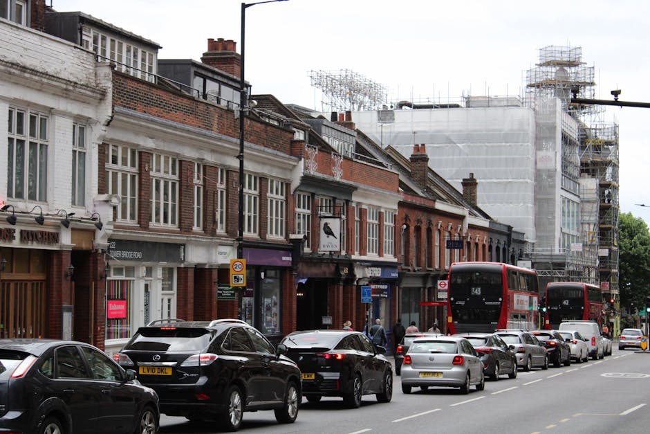 A street scene in Croydon, showing a row of traditional brick terraced houses with white window frames and pitched roofs, some with scaffolding and construction material indicating ongoing building work. In the foreground, a line of parked cars includes hatchbacks, sedans, and SUVs, with license plates visible. Two red double-decker buses are moving along the road, one numbered 343 and the other 42, suggesting urban transportation. Pedestrians are walking on the pavement, passing storefronts that feature shop windows and signage. The street appears busy with traffic and activity typical of Croydon’s residential and commercial area, providing context for home relocation or furniture transport activities supported by Man With a Van Croydon, which specializes in removals and moving services relevant to Croydon and Thornton Heath narrow streets.