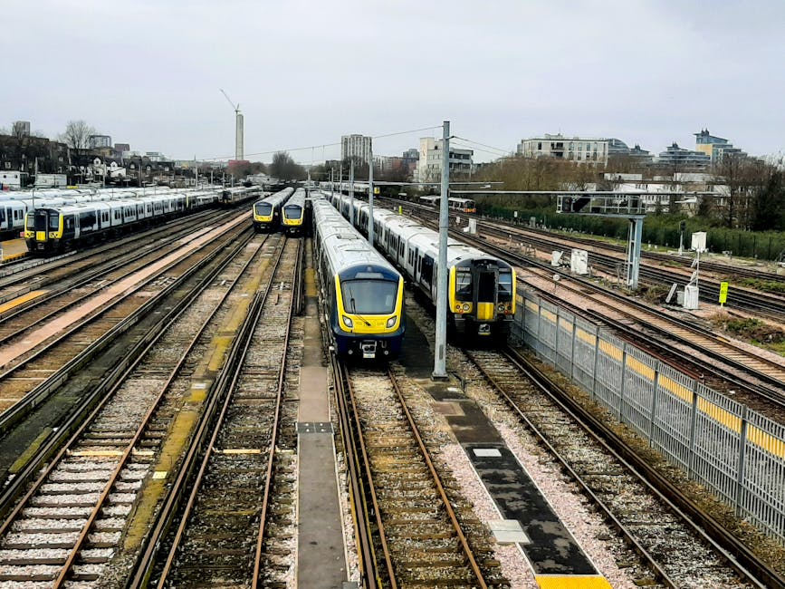 A view of multiple modern train carriages lined up at East Croydon station, with two yellow and grey passenger trains positioned on adjacent tracks, ready for departure or arrival. The scene is captured from an elevated perspective, showing several sets of railway lines extending into the distance, parallel to each other and separated by gravel beds. Overhead power lines are supported by tall metal poles that run alongside the tracks. In the background, there are urban buildings, including high-rise and mid-rise structures, with some trees and greenery visible further away. The sky is overcast, providing diffuse lighting across the scene. This railway environment, with its multiple tracks and urban surroundings, is relevant to the context of house removals and furniture transport, as transportation logistics often intersect with railway infrastructure for efficient moving services, such as those offered by Man With a Van Croydon.