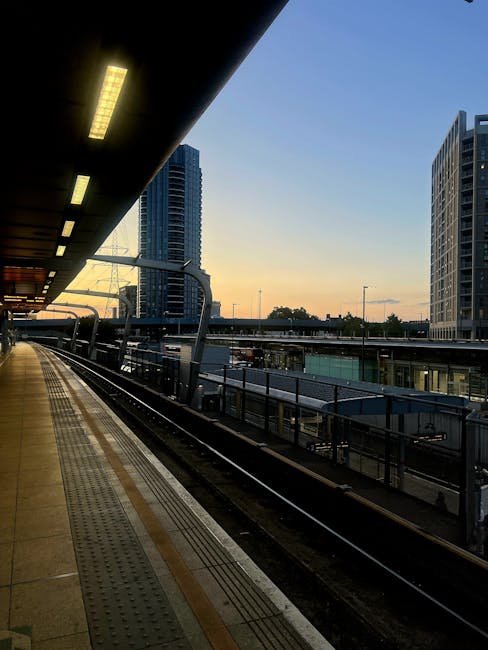 A view of East Croydon station platform during sunset, with overhead lighting illuminating the edge of the platform and empty train tracks visible below. Tall modern high-rise buildings line the background, and the sky displays a gradient of warm colors as evening approaches. The platform appears clean, with tactile paving and safety railings along the edge, indicative of a well-maintained area suitable for home relocation and furniture transport services in Croydon. The image captures the urban environment and transportation infrastructure relevant to moving logistics providers like Man With a Van Croydon.