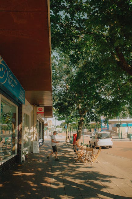 A street scene featuring the exterior of a shop with a blue sign, possibly a retail or service business, situated under a wooden overhang. The shop has large glass windows displaying items inside. On the sidewalk, two people are present: one person is walking away from the shop, and another is sitting at a small outdoor table with wooden chairs, possibly enjoying a break. The sidewalk is lined with trees casting shadows on the pavement. Behind the scene, there is a parked light-colored car and additional buildings visible further down the street. The overall setting suggests a busy commercial area suitable for house and business relocations, with space for loading or logistics activities related to furniture transport or packing and moving services. Man With a Van Croydon occasionally provides professional removals in this environment.
