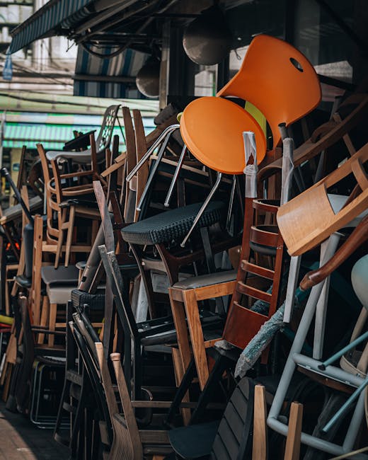 A dense stack of various used furniture items including wooden chairs, plastic chairs, and stools, stored indoors in what appears to be a warehouse or storage area. Some chairs are stacked on top of each other, with a mix of black, brown, and orange finishes. Several chairs have fabric or padded seats, while others are entirely wooden or plastic. The furniture is arranged in a haphazard manner, indicating a collection awaiting moving or disposal. The background shows a dark ceiling with beams and some industrial elements, and natural light filters into the space. This setting reflects the collection of household or commercial furnishings that might be involved in packing and home relocation services handled by Man With a Van Croydon, especially for clients seeking furniture transport or clearance in Croydon area for Purley Way shops and businesses.