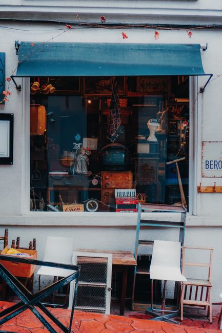 A street scene featuring the exterior of a shop with a blue sign, possibly a retail or service business, situated under a wooden overhang. The shop has large glass windows displaying items inside. On the sidewalk, two people are present: one person is walking away from the shop, and another is sitting at a small outdoor table with wooden chairs, possibly enjoying a break. The sidewalk is lined with trees casting shadows on the pavement. Behind the scene, there is a parked light-colored car and additional buildings visible further down the street. The overall setting suggests a busy commercial area suitable for house and business relocations, with space for loading or logistics activities related to furniture transport or packing and moving services. Man With a Van Croydon occasionally provides professional removals in this environment.