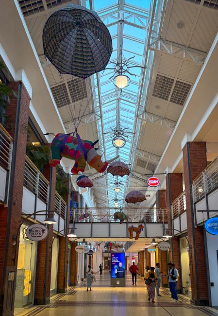 Inside the Croydon Whitgift Centre shopping mall, a high glass ceiling with a metal framework allows natural light to illuminate the corridor. Hanging from the ceiling are colorful decorative umbrellas and a large umbrella-shaped canopy, along with various plush toys and fabric umbrellas, creating a festive atmosphere. On the ground level, shoppers are visible walking along the tiled walkway, some carrying shopping bags, while others are standing and engaging in conversation. The mall features brickwork and metal railings along the upper level, with several storefronts, including a visible bakery shop called 'KoKo Bongo' to the left. The overall scene captures a busy indoor shopping environment, illustrating the typical setting where furniture or packing materials might be moved during house removals or home relocation logistics involving professional services like those provided by Man With a Van Croydon.