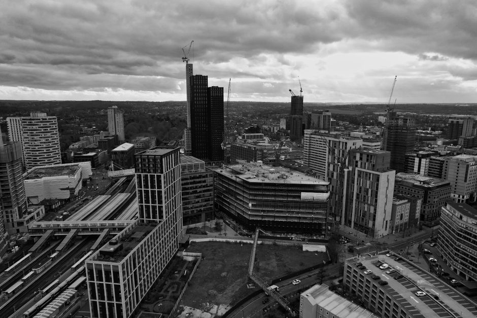 A black and white aerial photograph of a cityscape featuring multiple modern high-rise buildings, some of which are under construction with cranes visible on top. The scene includes a mix of commercial and residential structures, with a multi-level transportation hub or train station with elevated tracks at the lower left. Surrounding streets are busy with traffic and parked vehicles. In the foreground, a large green space is visible, possibly part of a recent development or open area. The overcast sky creates a moody atmosphere. This urban environment depicts a developing area suitable for home relocation or furniture transport, with ongoing construction projects that may require professional moving services, such as those offered by Man With a Van Croydon, especially around busy city centre locations like Croydon.